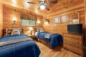 Ein schlicht eingerichtetes Schlafzimmer im Blockhausstil mit zwei Betten, einem Schminktisch, einem Fernseher und einem Deckenventilator.