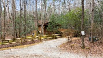 A rustic cabin with a wooden deck and a covered area, surrounded by tall trees and a gravel path.