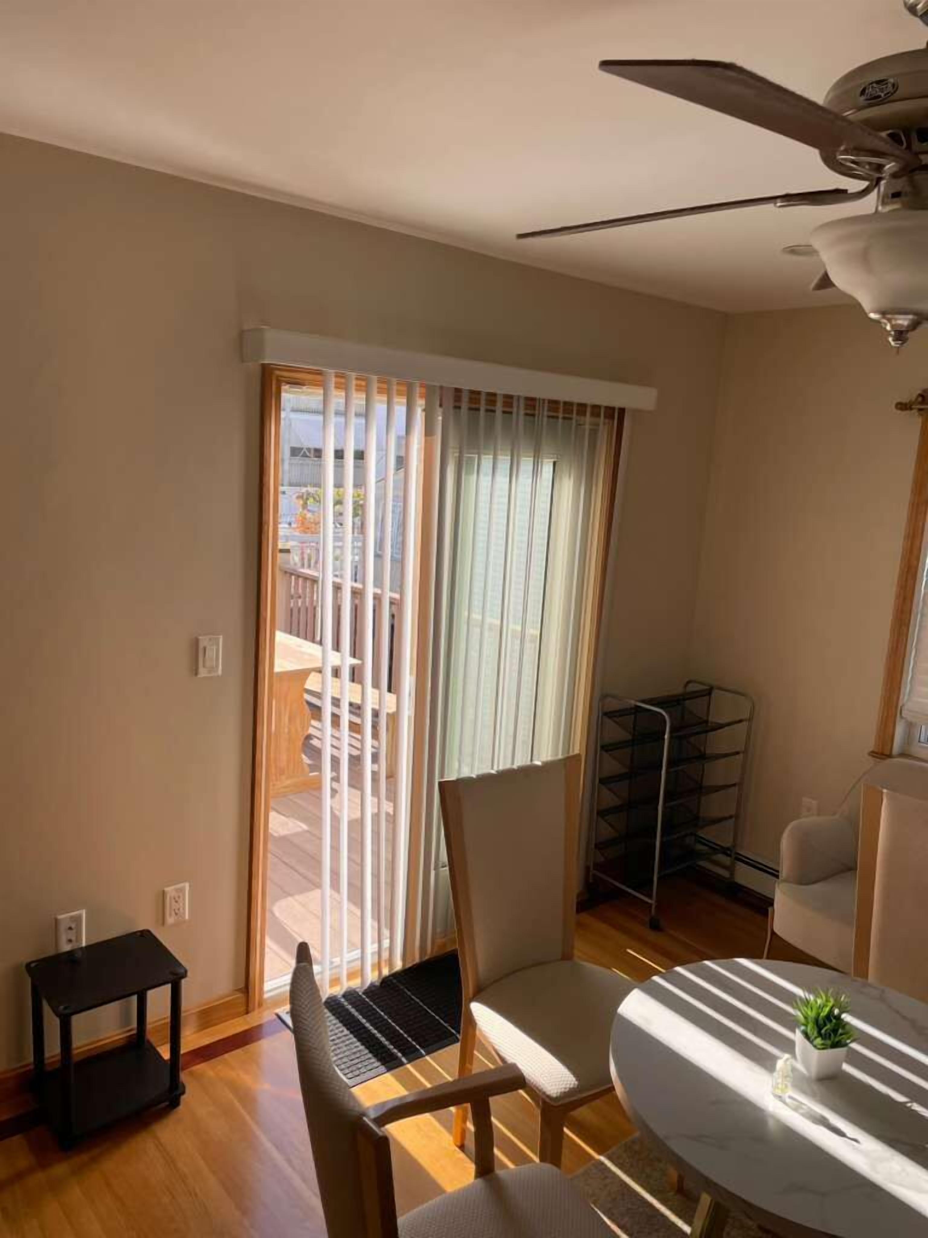 Una stanza con ventilatore a soffitto, un tavolo da pranzo con sedie, un piccolo tavolino e vista su una zona esterna con balcone tramite una porta scorrevole in vetro.