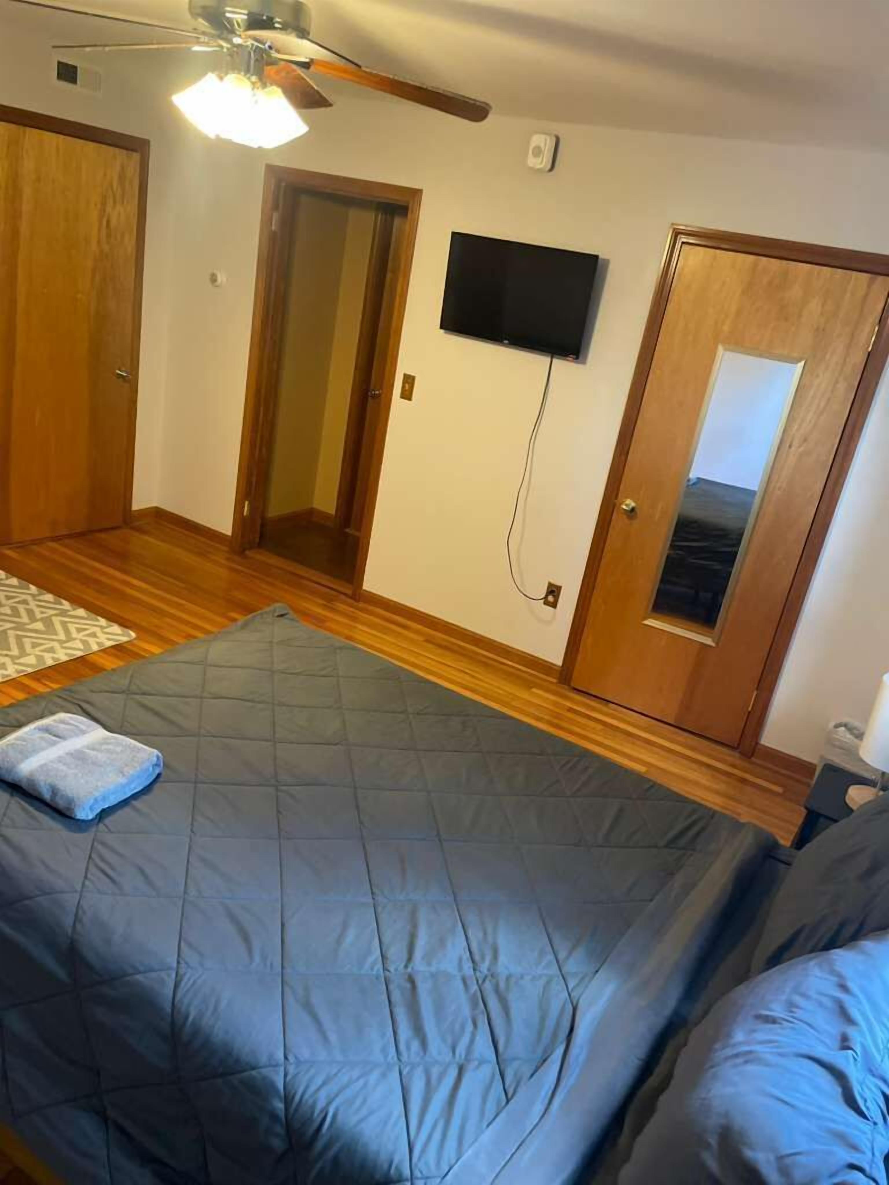 A bedroom with a bed, a ceiling fan, a TV mounted on the wall, and wooden doors.