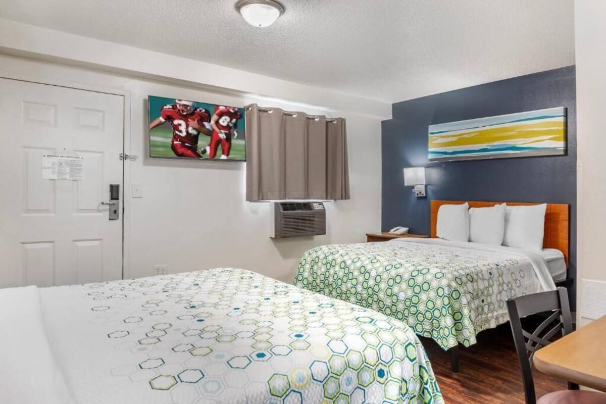 Habitación de hotel con dos camas, un escritorio, una silla, un cuadro, una ventana con cortinas y aire acondicionado de pared.