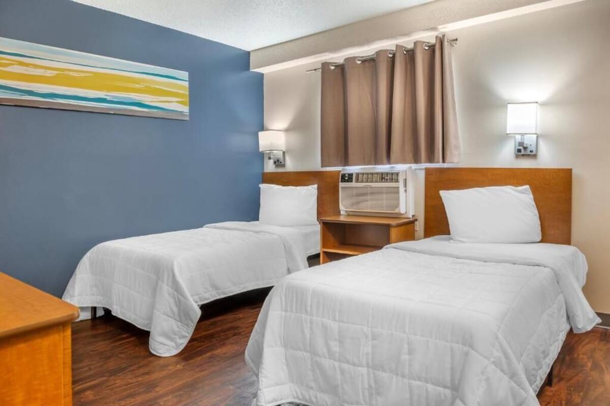 Habitación de hotel con dos camas individuales, una mesita de noche de madera, un aire acondicionado instalado en la pared y un cuadro colgado.