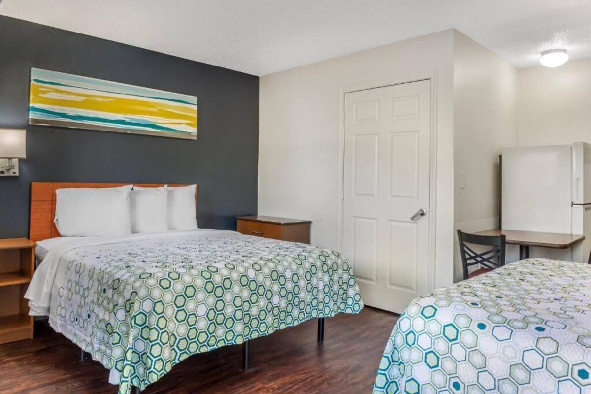 Habitación de hotel con cama, escritorio y silla. Se observa una puerta y un cuadro en la pared.
