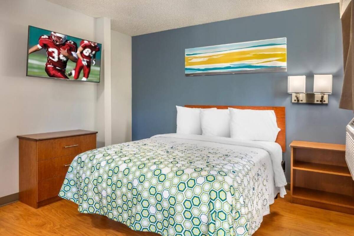Una habitación de hotel con una cama, una mesita de noche de madera, un televisor en la pared y un cuadro sobre la cama.