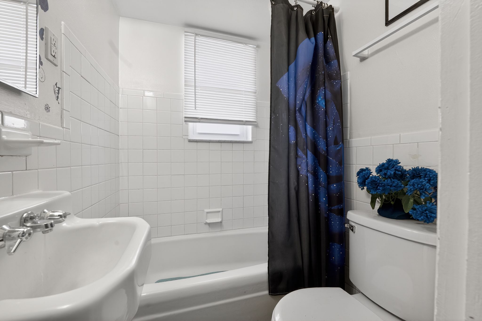 A bathroom with a white sink, a bathtub, and a toilet. There is a window with blinds, a shower curtain with a starry pattern, and a potted plant with blue flowers on the toilet tank.