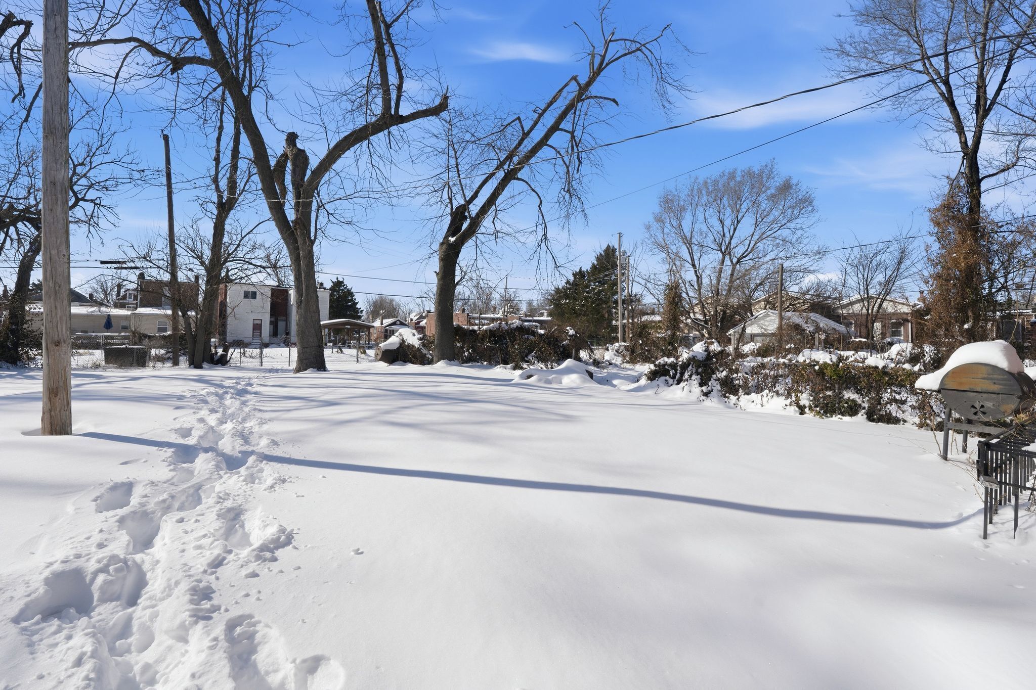 Un quartier résidentiel enneigé, avec des arbres sans feuilles, un ciel dégagé et des maisons en arrière-plan.