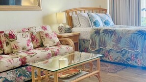 Ett sovrum med en blommönstrad soffa, ett soffbord i glas, en säng i trä med ett påslakan med lövmönster, en sänglampa och en inramad tavla på väggen.