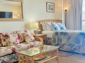 A bedroom with a floral sofa, a glass coffee table, a wooden bed with a leaf-patterned duvet, a bedside lamp, and a framed picture on the wall. - Stylish Studio- Stunning City View - Parking (Honolulu)