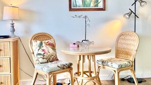 Ett rum med två fåtöljer i rotting, ett runt bord, en inramad botanisk tavla på väggen, en golvlampa och ett litet sidobord med en lampa.