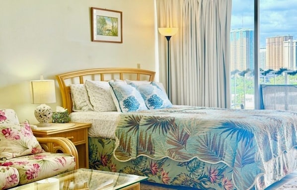 A hotel room with a bed, a nightstand, a lamp, a floral sofa, and a view of buildings. - Stylish Studio- Stunning City View - Parking (Honolulu)