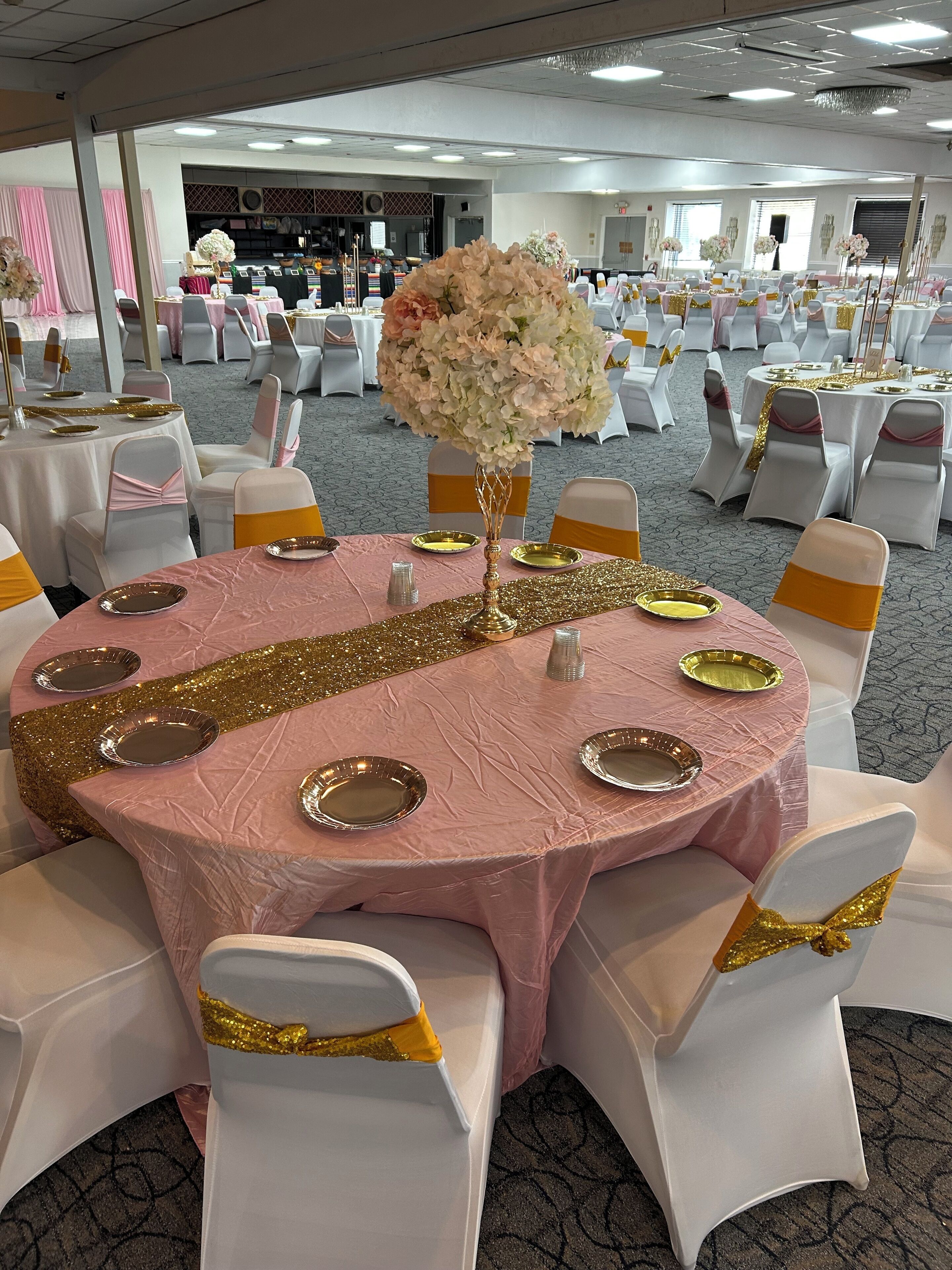 A banquet hall with round tables covered in pink tablecloths, white chairs with gold sashes, and gold glittery borders.