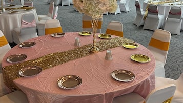 Une salle de banquet avec des tables rondes recouvertes de nappes roses, des chaises blanches ornées de rubans dorés et des bordures scintillantes dorées.