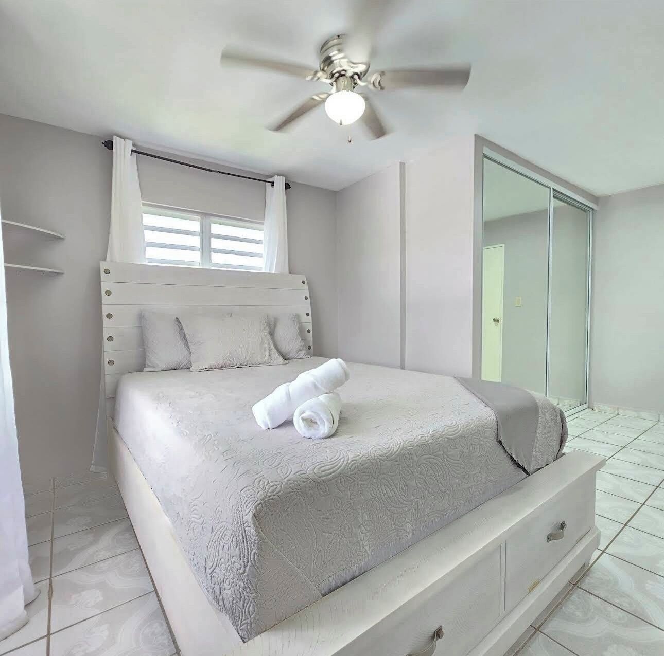 Un dormitorio con cama, ventilador de techo, ventana con cortinas y puerta corrediza de vidrio.