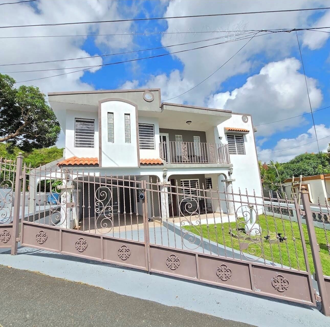 Una casa de dos pisos con balcón, entrada protegida por un portón y césped bien cuidado.