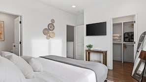 Une chambre avec un lit, un téléviseur fixé au mur, une petite table avec une plante et une armoire contenant des vêtements et un ventilateur.