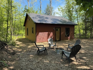 A small cabin with a red roof and yellow siding, surrounded by trees. There are two Adirondack chairs and a metal fire pit in front. - Secluded cabin on the trails- cabin #1 (Petoskey)