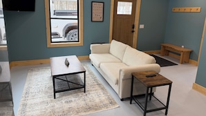 A living room with a beige sofa, a coffee table, a small wooden table, a bench, and a TV mounted on the wall. - Secluded cabin on the trails- cabin #1 (Petoskey)