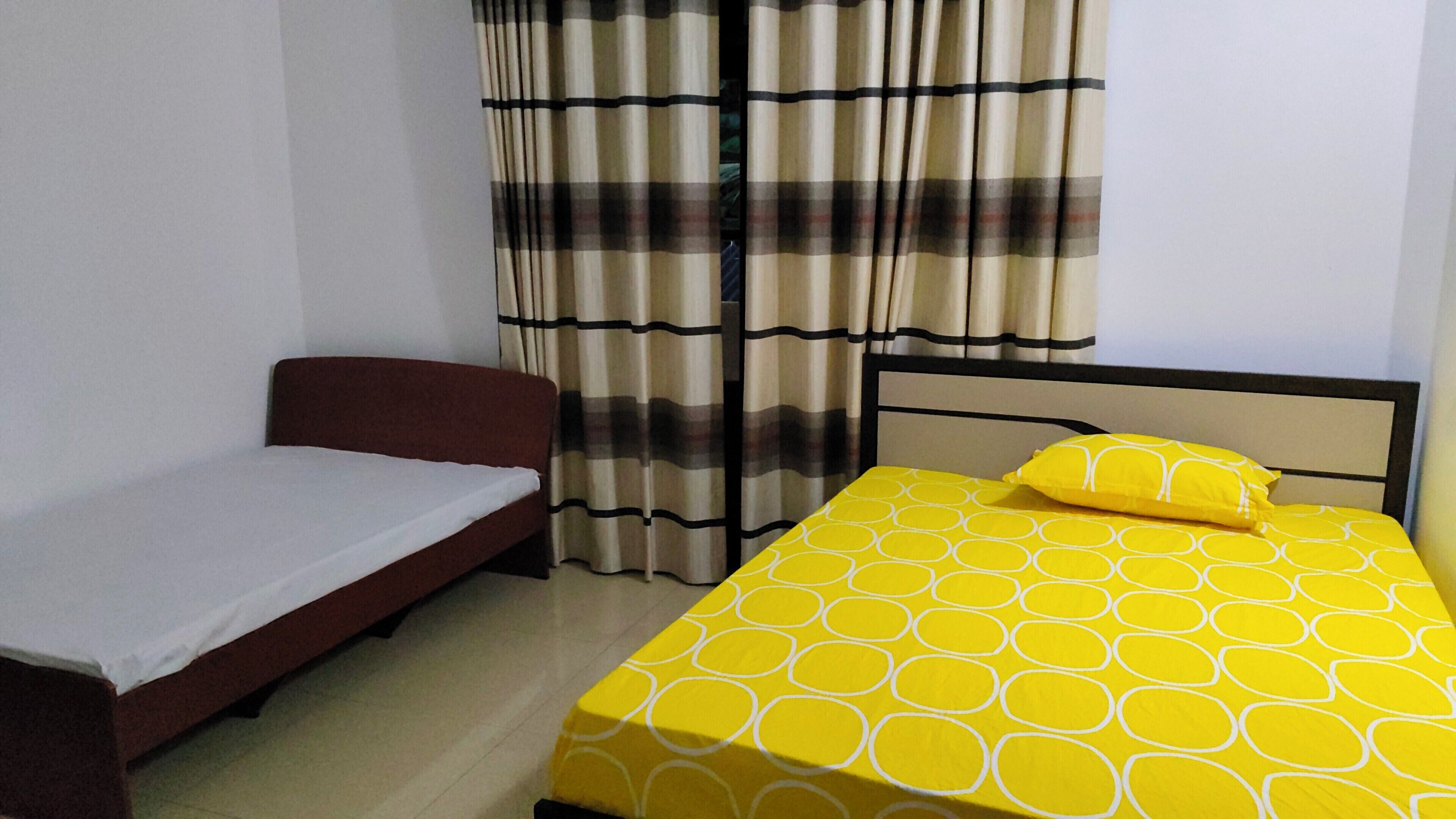 Une chambre avec un lit simple recouvert d’une couvre-lit jaune à motifs et agrémentée de deux oreillers, un plus petit lit avec un drap blanc, et une fenêtre ornée de rideaux à carreaux.