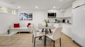 Ein modernes Wohnzimmer mit einem weißen Sofa, einem roten Kissen und einem Couchtisch. Es gibt eine Essecke mit einem runden Tisch und Stühlen. Die Küche verfügt über weiße Schränke und eine Mikrowelle.