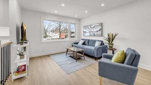 Una sala de estar moderna con un sofá gris, una mesa de centro y un televisor sobre un mueble bajo. Hay una ventana con vistas a árboles y un camión afuera.