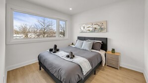 Un dormitorio con un ventanal grande, una cama con ropa de cama gris, una mesita de noche con una planta y un cuadro en la pared.