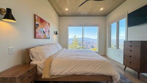 A bedroom with a bed, a dresser, a painting, and a large window with a mountain view. - Luxury Cabin w/ 180 Views, Hot Tub & Firepit Near Dollywood & Nat'l Park Bird's-eye View by Avantstay (Sevierville)