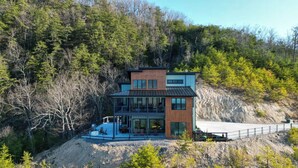 A modern two-story house with a large glass facade, situated on a hillside with a view of a body of water. - Luxury Cabin w/ 180 Views, Hot Tub & Firepit Near Dollywood & Nat'l Park Bird's-eye View by Avantstay (Sevierville)