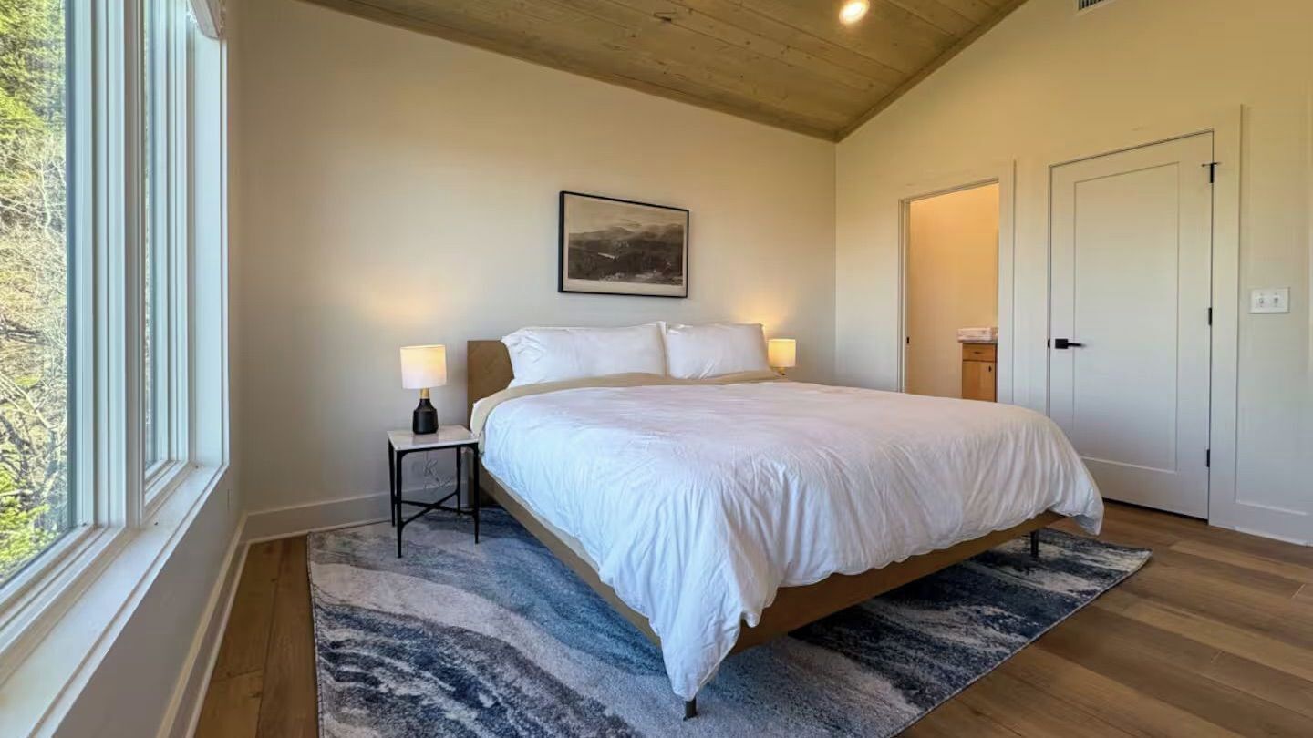 A bedroom with a bed, two bedside lamps, a rug, and a window.