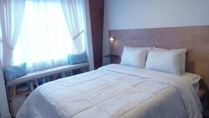 Ein Schlafzimmer mit Holzboden, einem großen Bett mit weißer Bettwäsche, einem Fenster mit transparenten Vorhängen und einem Nachttisch mit Lampe.