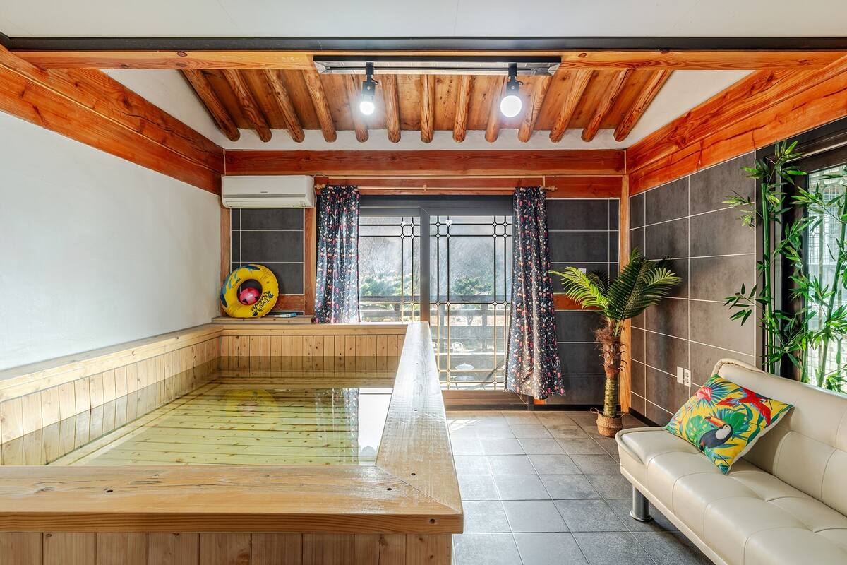 A wooden-floored indoor swimming pool with a bench, a sofa with a colorful pillow, and a potted plant.