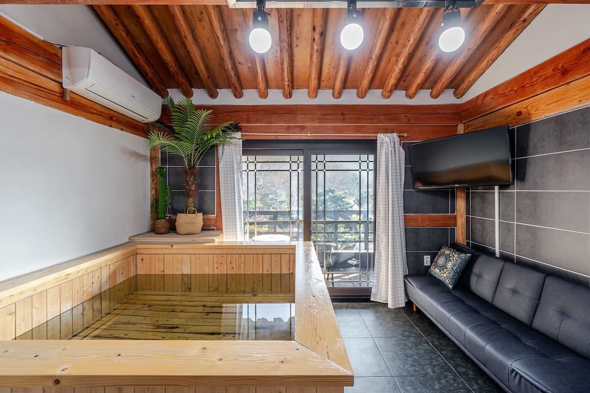 A sauna room with a wooden bench, a black leather sofa, a TV mounted on the wall, and a large window with curtains.