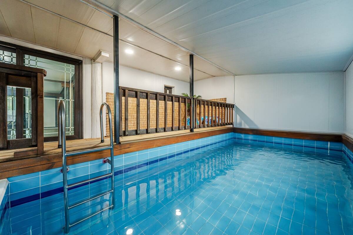 An indoor swimming pool with clear blue tiles, a wooden deck, and a metal ladder.