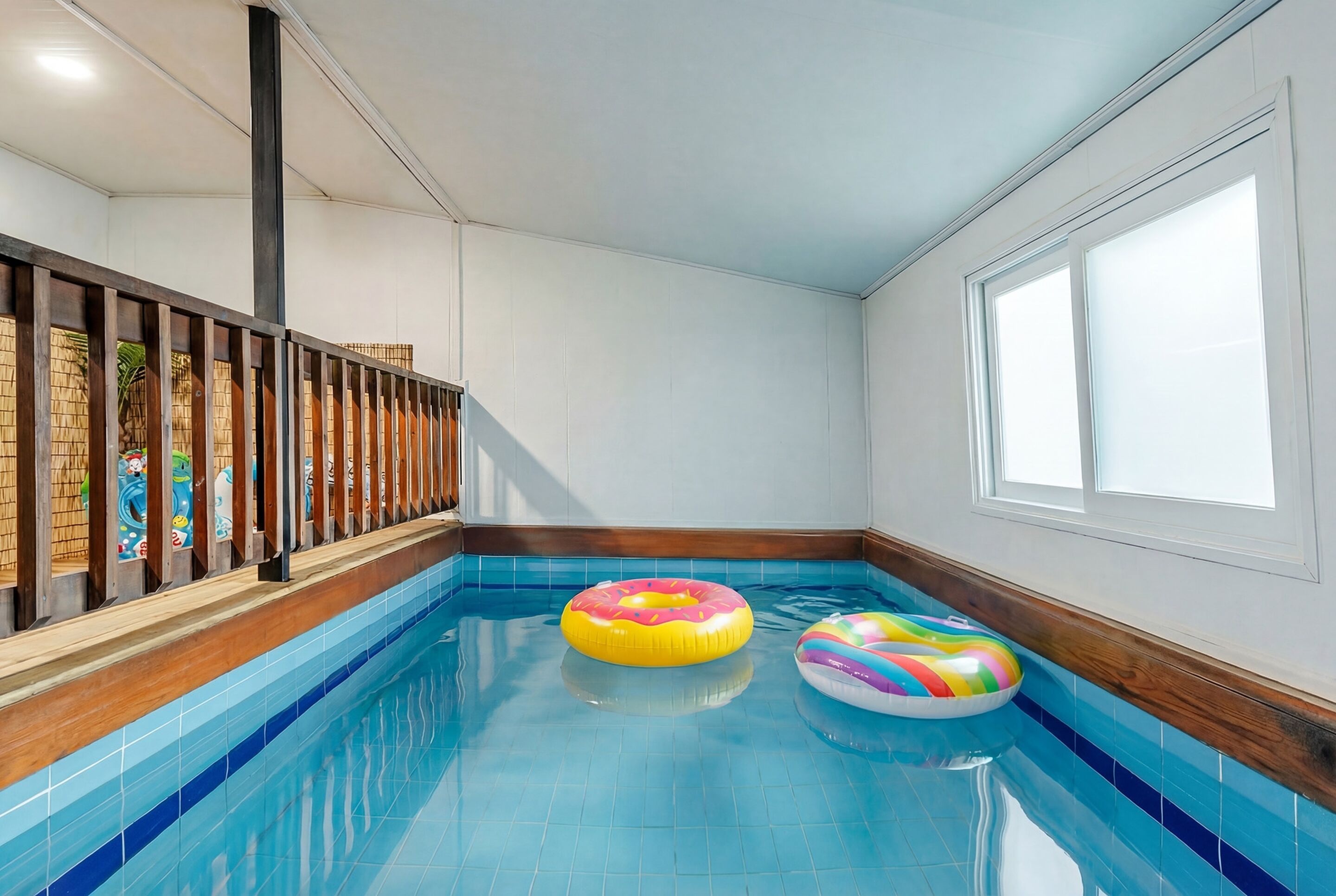 Una piscina interior con dos flotadores de colores, paneles de madera y un ventanal grande.