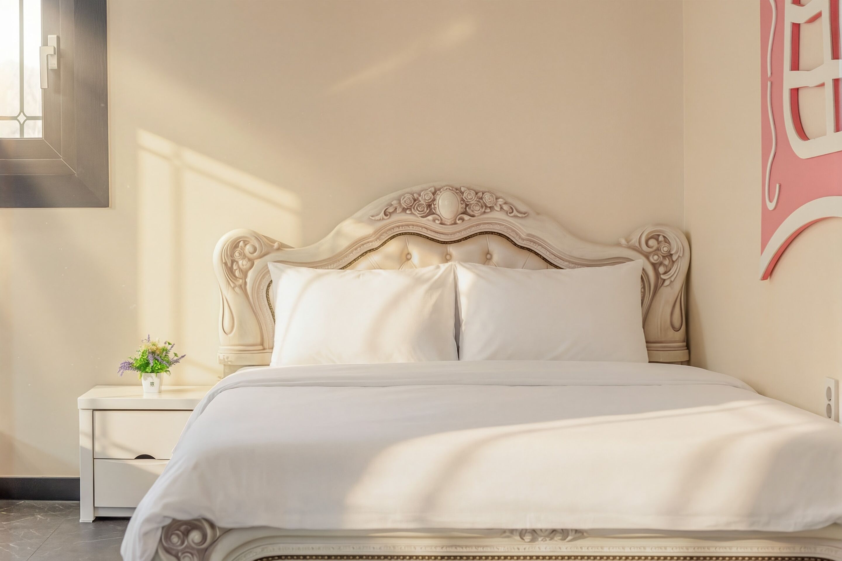A neatly made bed with a classic headboard, a bedside table with a vase of flowers, and a wall-mounted artwork.
