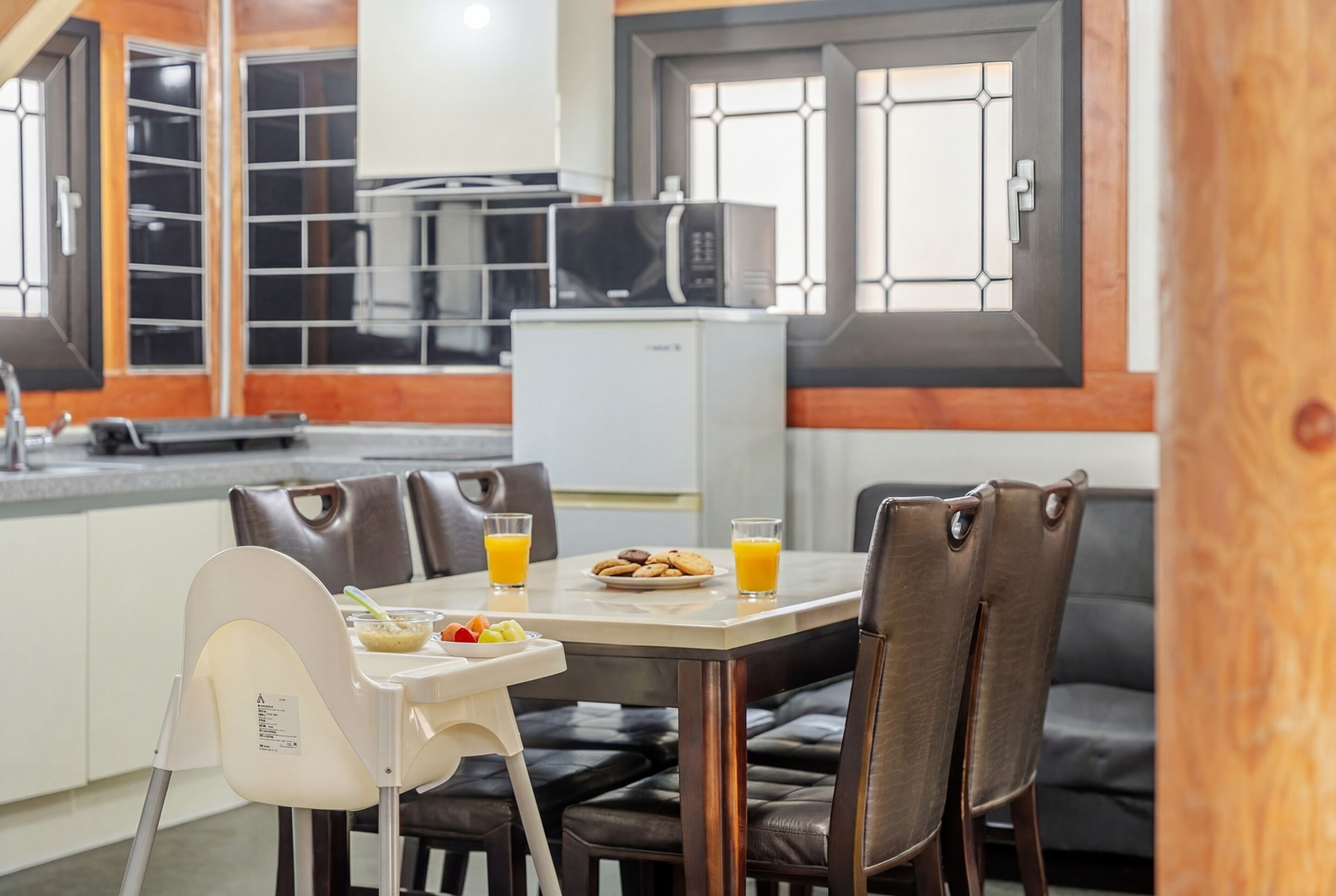 A modern kitchen with a dining area, featuring a microwave, refrigerator, and a table set for breakfast with orange juice and cookies.