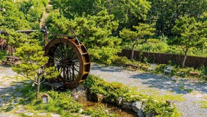 A traditional water wheel in a garden setting with stone pathways, small wooden structures, and lush greenery. - Gyeongju Palace Hanok Kids Pool Villa (Gyeongju)
