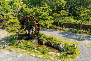 Une roue à eau traditionnelle, installée dans un jardin agrémenté de chemins de pierre, de petites constructions en bois et d’une végétation luxuriante.