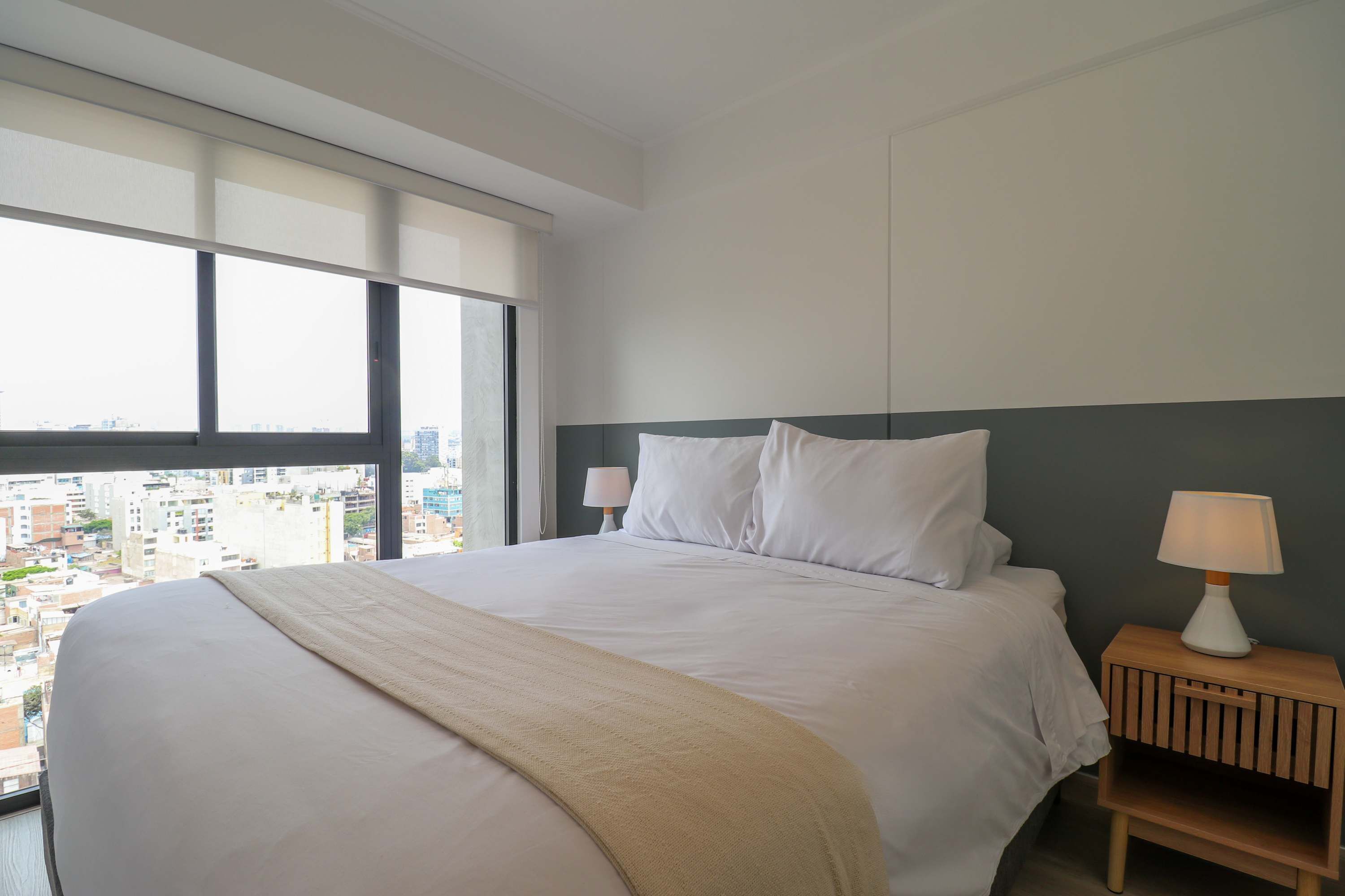 Ein Hotelzimmer mit einem großen Bett, einem Nachttisch mit Lampe und Blick auf die Stadt durch große Fenster.