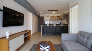 Un salon moderne avec un canapé gris, une table basse en bois sur laquelle sont posés des livres, un téléviseur à écran plat fixé au mur et un coin cuisine équipé de tabourets de bar et d’un éclairage suspendu.