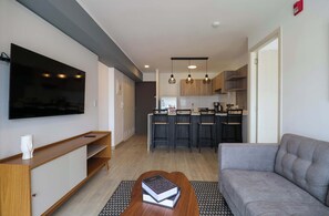 A modern living room with a grey sofa, a wooden coffee table with books, a flat-screen TV mounted on the wall, and a kitchen area with bar stools and pendant lighting. - w* | Engaging 2BR in the Heart of Barranco (Barranco)