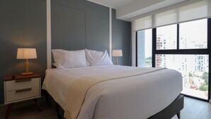 Une chambre d’hôtel moderne avec un grand lit, deux tables de chevet avec des lampes, une vue sur la ville grâce à de grandes fenêtres, et un panorama urbain visible à l’extérieur.