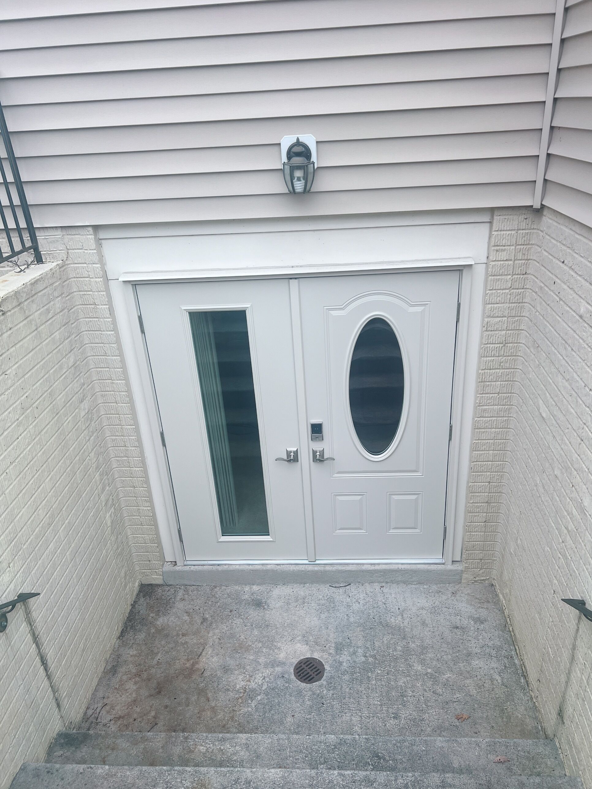 A white double door with a glass panel and a peephole, set in a concrete stairwell with a metal handrail and a vent.