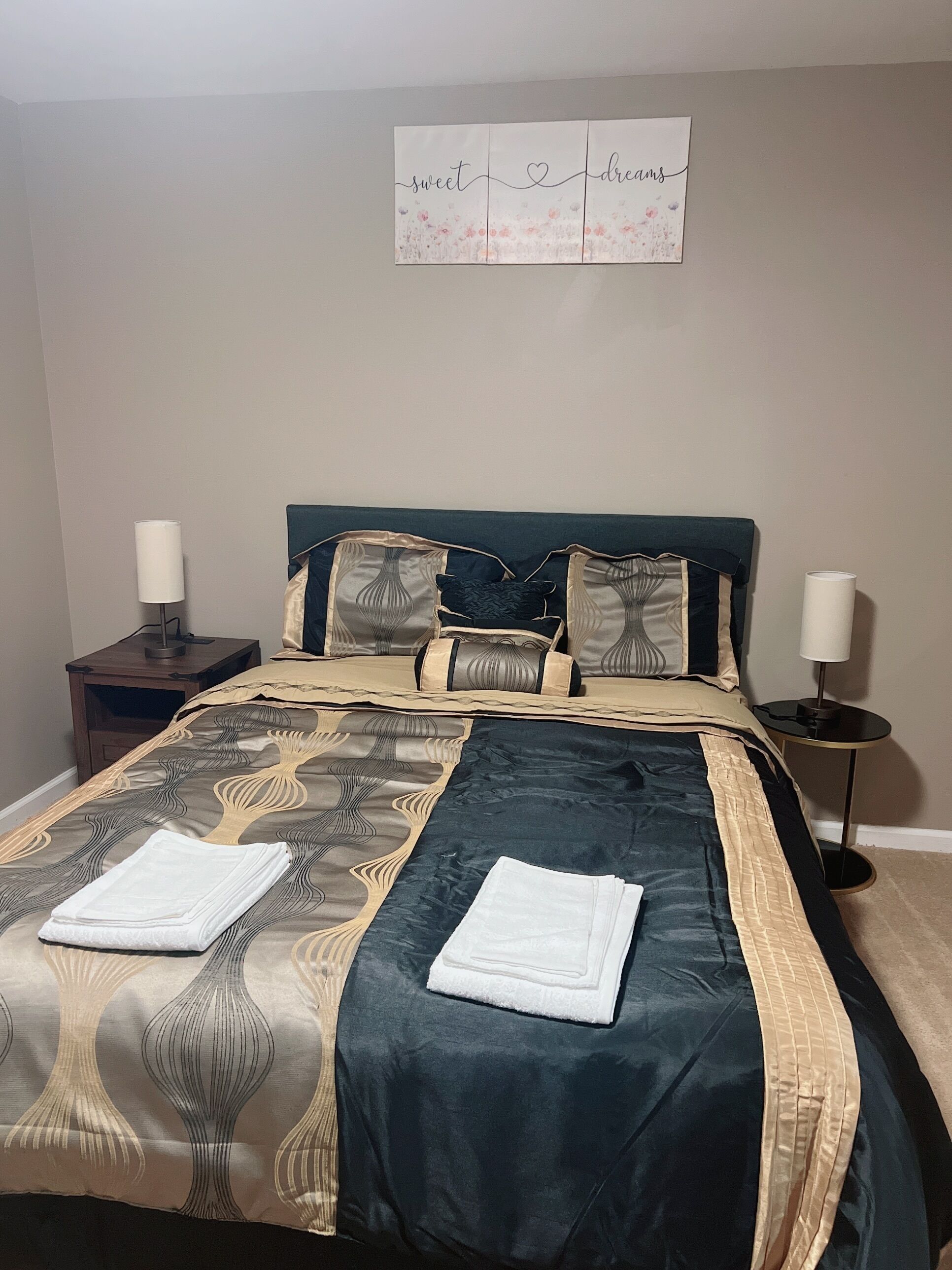 Una cama bien hecha con una colcha estampada y almohadones, dos mesitas de noche con lámparas, y una decoración en la pared sobre la cama.