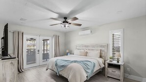 Ein Schlafzimmer mit einem Bett, einem Deckenventilator, einer Klimaanlage, einem Nachttisch, einer Lampe und einem Fenster mit Jalousien.