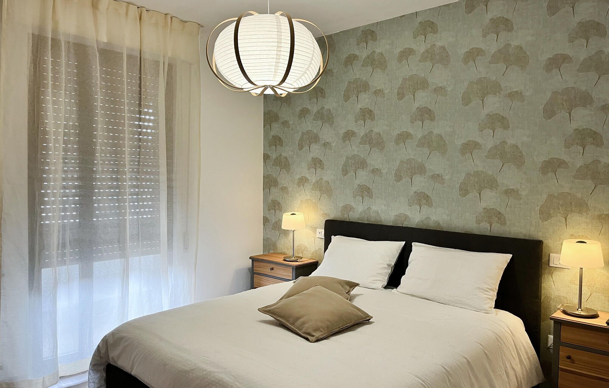 A bedroom with a bed, two bedside tables, a lamp, a window with sheer curtains, and a wall with a leaf pattern.