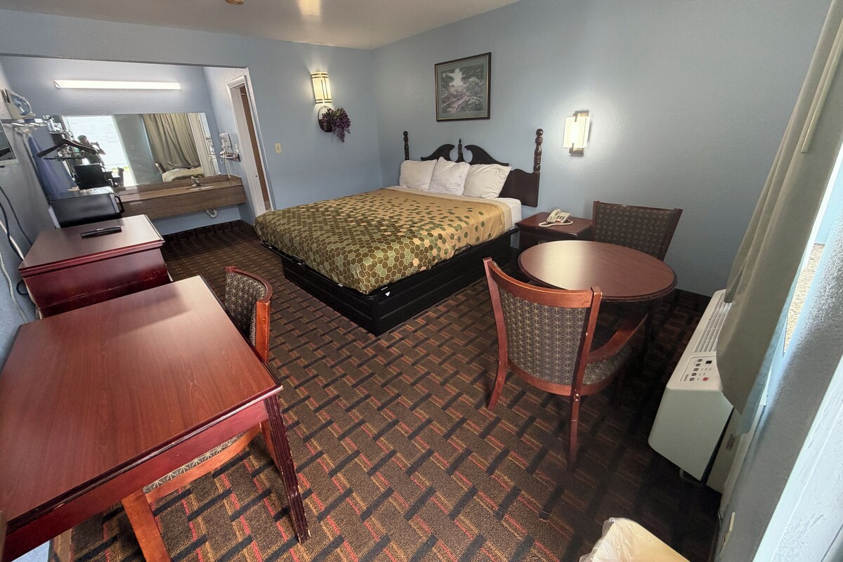 A hotel room with a bed, a desk, a chair, a table, a telephone, and a radiator.