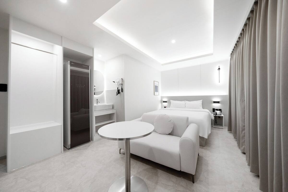 Ein modernes Hotelzimmer mit einem weißen Sofa, zwei Betten, einem runden Tisch, einem Badezimmer mit Spiegel und einem Kleiderschrank.