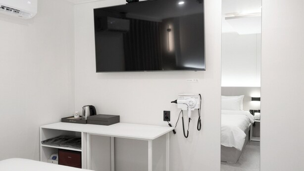 A modern hotel room with a flat-screen TV mounted on the wall, a desk with a kettle and a coffee maker, a hairdryer, and a bed with white bedding. - Bucheon Songnae Number 25 (Bucheon)