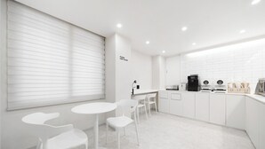 A modern kitchen with white cabinetry, a black coffee machine, and a small dining area with white chairs and tables. - Bucheon Songnae Number 25  (Bucheon)