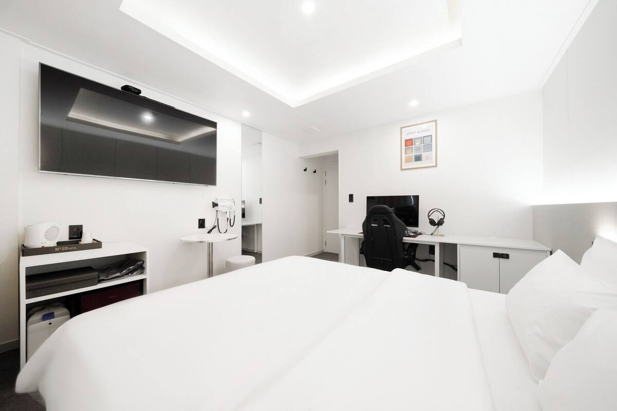 Ein modernes Hotelzimmer mit einem großen Bett, einem Schreibtisch mit Stuhl, einem Flachbildfernseher, einem Spiegel und einem kleinen Tisch mit Lampe.